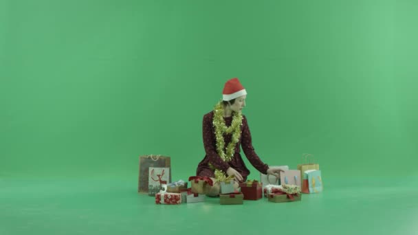 Una joven está sentada y buscando regalos de Navidad cerca de ella en la pantalla verde — Vídeos de Stock