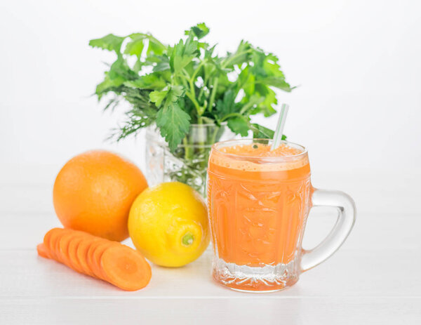 Carrot smoothies, a bunch of greenery, lemons and an orange on a wooden table.