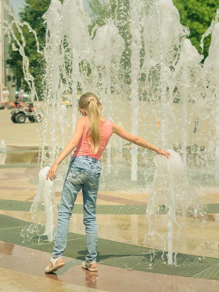 Girl in jeans plays with the water jets in the fountain.