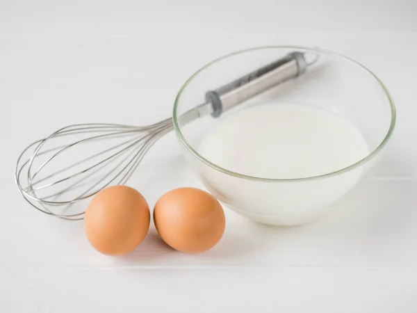 Dos huevos, un batidor y un tazón de leche sobre una mesa de madera blanca . — Foto de Stock