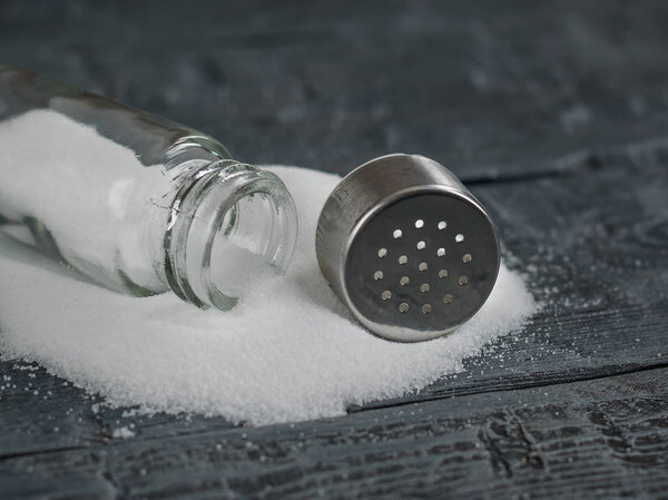 Белая соль, выливаемая из открытой солонки на деревянный стол
.