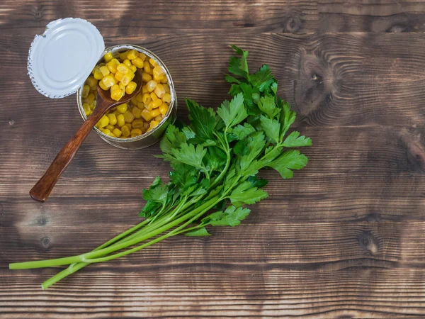 Canned corn in an open aluminum cans and parsley on a dark wooden table. The view from the top.