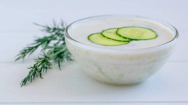 Salatalık dilimleri ve dereotu Beyaz ahşap bir masa üzerinde bir cam kase cacık.