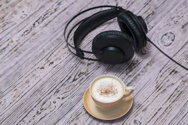 A Cup of coffee and black headphones with a wire on a wooden table.