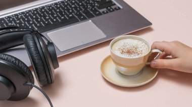 Elinde bir fincan kahve, dizüstü bilgisayar ve pembe arka planda kulaklık olan bir el. Görev.