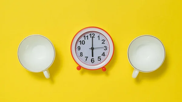 ถ้วยกาแฟสองถ้วยและนาฬิกาปลุกสีแดงบนพื้นหลังสีเหลือง . — ภาพถ่ายสต็อก