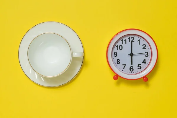 Eine Kaffeetasse mit Untertasse und ein roter Wecker auf gelbem Hintergrund. flache Lage. — Stockfoto