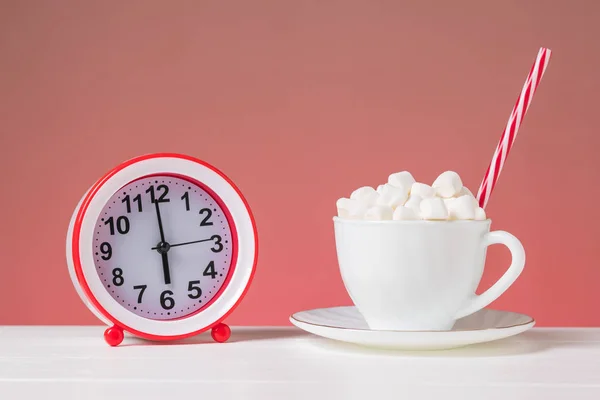 Una taza blanca llena de malvavisco y un despertador rojo sobre una mesa blanca sobre un fondo rosa . — Foto de Stock