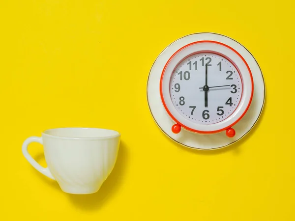 Чашка белого кофе и будильник на блюдце на желтом фоне. Плоский лежал . — стоковое фото