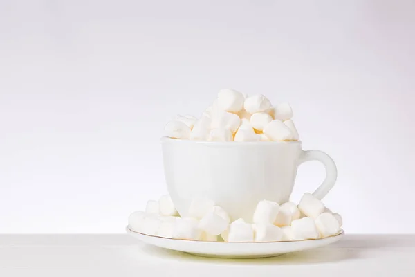 Pequeños trozos de malvavisco en una taza blanca y platillo sobre una mesa blanca . — Foto de Stock