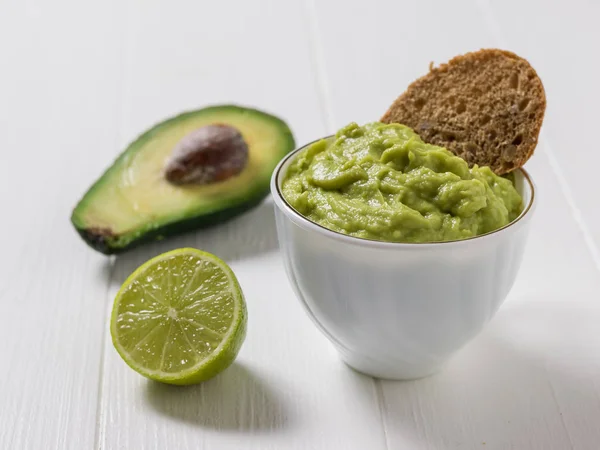 Гуакамоле с куском хлеба в белой миске, половиной авокадо и половиной лайма. Мексиканская вегетарианская еда авокадо . — стоковое фото