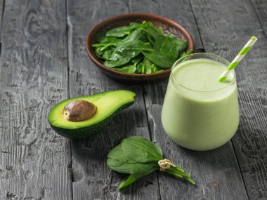 A bowl of spinach leaves, half an avocado, and a glass of smoothies on a black wooden table. Fitness product.