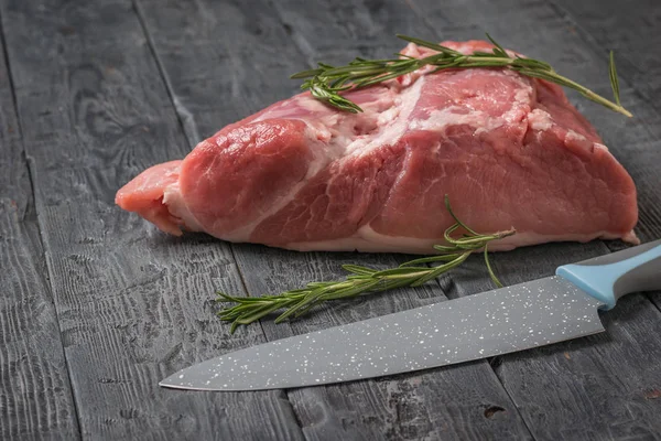 Um grande pedaço de porco com alecrim e uma faca em uma mesa de madeira preta . — Fotografia de Stock