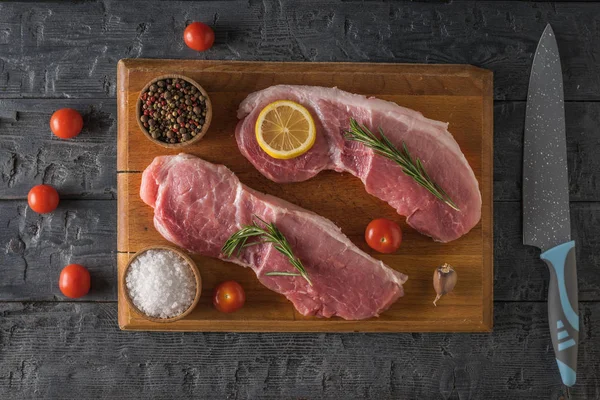 Dos filetes de cerdo en una tabla de cortar con especias y tomates en una mesa de madera . —  Fotos de Stock
