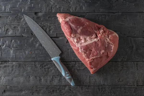 A piece of pork with a large knife on a black wooden table. The view from the top. — ストック写真