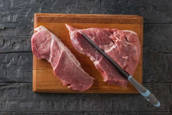 Uma faca corta um grande pedaço de porco em uma tábua de corte em uma mesa de madeira. A vista de cima . — Fotografia de Stock
