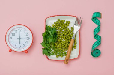 Alarm clock, measuring tape and a plate with green peas on a pink background.