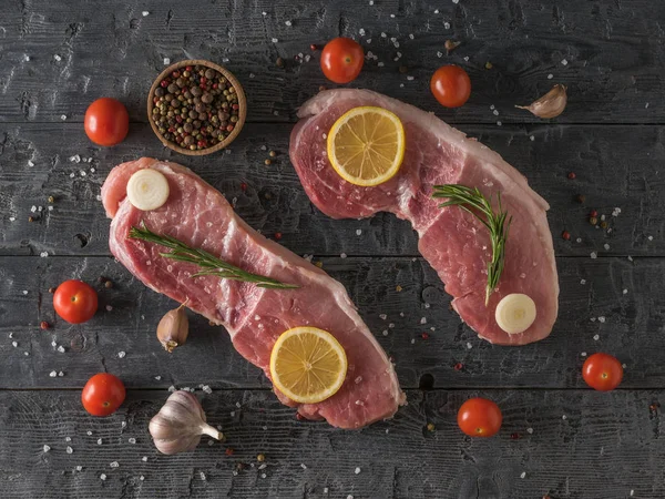 Two large pork steaks with lemon, tomatoes, spices and garlic on a wooden table. The view from the top. — Stock fotografie