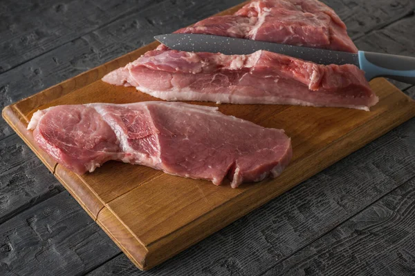 Un cuchillo grande corta filetes de un pedazo grande de cerdo en una tabla de cortar . —  Fotos de Stock