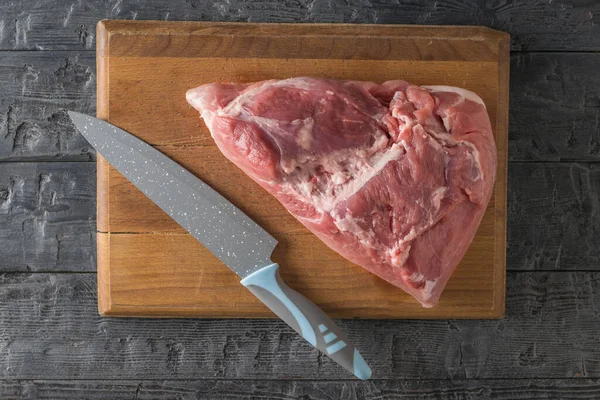 Um grande pedaço de carne de porco em uma tábua de corte e uma faca grande em uma mesa de madeira. A vista de cima . — Fotografia de Stock