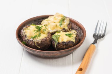 Fork near a clay bowl with baked mushrooms on a white wooden table.