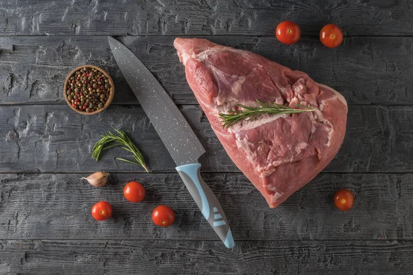 A large blue knife, a piece of pork , and cherry tomatoes on a wooden table. The view from the top. — Stock Fotó