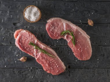 A wooden bowl with coarse sea salt and two pork steaks on a black wooden table. The view from the top.