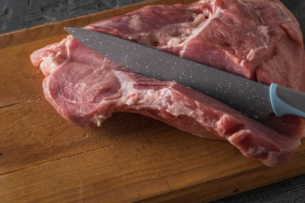 En kniv skærer en svinekød bøf fra et stort stykke kød på et træbord . - Stock-foto