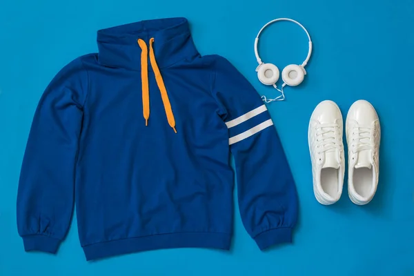 Sweatshirt, vita sneakers och vita hörlurar på en blå bakgrund. — Stockfoto