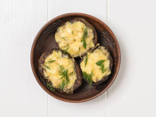 Three large baked mushrooms in a clay bowl on a white wooden table. The view from the top.