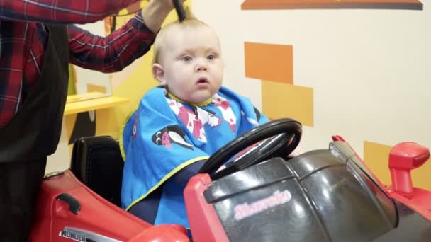 El niño corta al barbero. Se sienta en una silla que parece un coche . — Vídeos de Stock