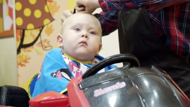 Le petit garçon coupe le coiffeur. Il est assis sur une chaise qui ressemble à une voiture. . — Video