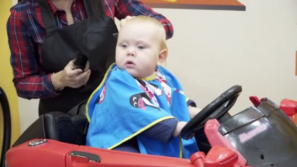 El niño corta al barbero. Se sienta en una silla que parece un coche . — Vídeos de Stock