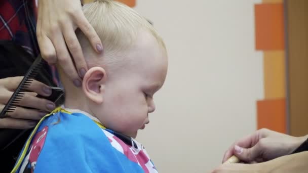 El niño corta al barbero. Se sienta en una silla que parece un coche. Mamá, distrae y juega con él. Primer plano de la cabeza . — Vídeos de Stock