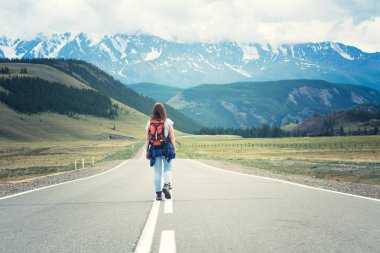 Lonely girl with a backpack walking along the road. In the distance, mountains. Altai, Russia. clipart