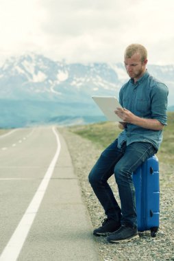 Yalnız adam bir bavul ve tablet vasıl bakarak oturur. Otobüs veya araba bekliyor. Yolun dağlarda.