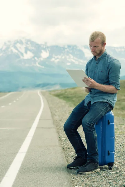 Einsamer Mann sitzt auf einem Koffer und blickt auf Tablet. Warten auf Bus oder Auto. die Straße in den Bergen. Stockbild