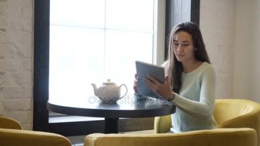 Genç esmer kız yakın pencere ve tablet pc içine bakmak masada oturur