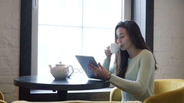 Chica morena joven se sienta en la mesa cerca de la ventana y mirar en la tableta PC. Beber té . — Vídeo de stock