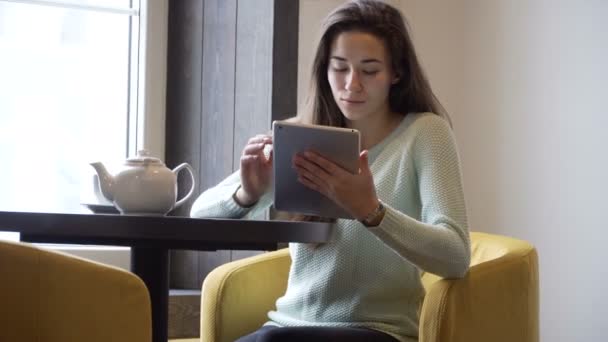 Jonge brunette meisje zit op de tafel in de buurt van venster en werken op tablet pc — Stockvideo