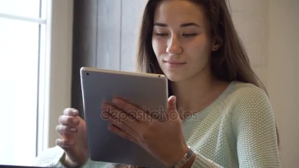 Jonge brunette meisje zit op de tafel in de buurt van venster en werken op tablet pc — Stockvideo