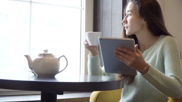 Menina senta-se a uma mesa e beber chá com um tablet pc em mãos — Vídeo de Stock