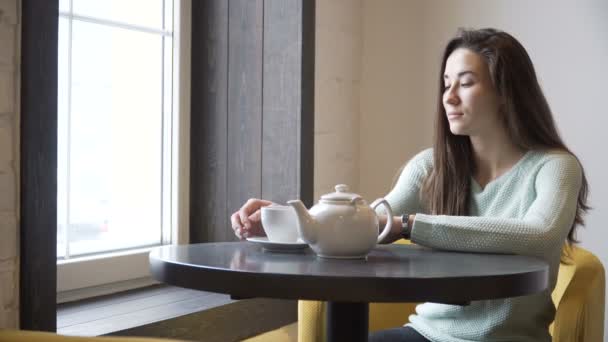 Menina bonita sentada a uma mesa e beber chá. Olhos sonhados a olhar pela janela . — Vídeo de Stock