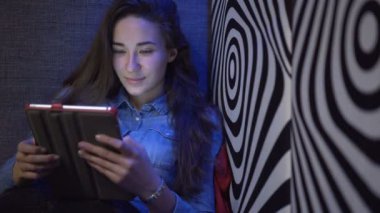 Tablet pc ile bir kanepede oturan genç güzel kız. Bir zebra gibi duvara. Çok renkli ışık. Kamera yavaş yavaş hareket eder. Elektronik Steadicam.
