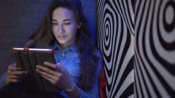 Mooi meisje zittend op een bank met een tablet-pc. Tegen de muur als een zebra. Multi gekleurd licht. De camera beweegt langzaam. Elektronische Steadicam. — Stockvideo