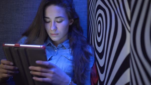 Ung smuk pige sidder på en sofa med en tablet-pc. Mod væggen som en zebra. Multi-farvet lys. Kameraet bevæger sig langsomt. Elektronisk Steadicam . – Stock-video