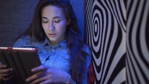 Krásná mladá dívka sedí na pohovce s tablet pc. Proti zdi jako zebra. Vícebarevné světlo. Kamera se pohybuje pomalu. Elektronické Steadicam. — Stock video