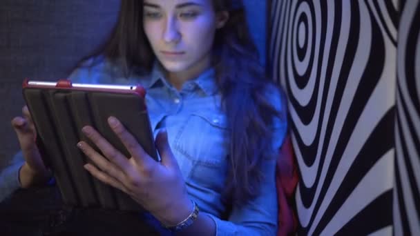 Όμορφη κοπέλα κάθεται σε έναν καναπέ με ένα tablet-pc. Ενάντια στον τοίχο, όπως μια ζέβρα. Πολύχρωμα light. Η κάμερα κινείται αργά. Ηλεκτρονική Steadicam. — Αρχείο Βίντεο
