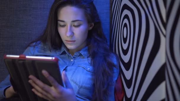 Menina bonita jovem sentada em um sofá com um tablet-pc. Contra a parede como uma zebra. Luz multicolorida. A câmara move-se devagar. Steadicam eletrônico . — Vídeo de Stock