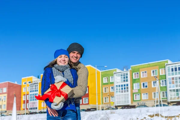 Mann und Mädchen umarmen sich auf einem Hintergrund farbiger Häuser mit Geschenk — Stockfoto